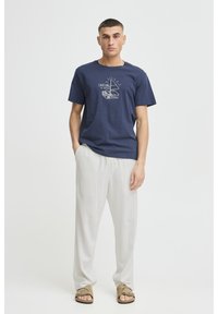 Námořnicky modré bavlněné tričko s bílým potiskem s motivem plachetnice; kombinováno s lehkými béžovými lněnými kalhotami a hnědými sandály.