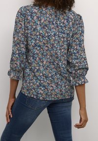 Kvinde med krøllet hår iført en blå blomstrede bluse med 3/4 ærmer og mørkeblå tætsiddende jeans, står foran en ensfarvet baggrund.