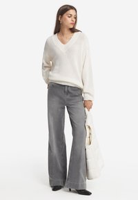 Vit stickad tröja med v-ringning, i kombination med ljusgrå utsvängda jeans. Modellen håller en vadderad vit väst och har på sig svarta klackskor.