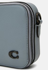 Ljusblå läderväska med en texturerad yta, som har en svart dragkedja och metallisk detalj. Kompakt rektangulär form, minimalistisk design.