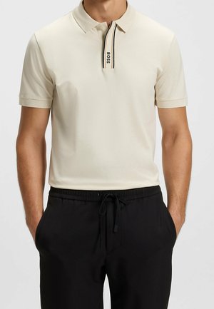 Mann trägt ein hellbeiges kurzärmeliges Poloshirt mit einem vertikalen BOSS-Logo-Streifen und schwarze Kordelhose, Hände in den Taschen.