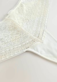 Lingerie en dentelle blanche avec des motifs floraux et des détails en maille sur un fond de tissu côtelé, exposée sur une surface claire.