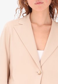 Blazer beige con chiusura a un bottone, revers a punte e texture liscia. La vestibilità su misura accentua la forma.