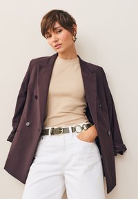 Dwurzędowa marynarka w ciemnym fioletowym kolorze, jasnobeżowy prążkowany top, białe dżinsy, oliwkowo-zielony pasek ze złotymi okrągłymi akcentami. Gładkie tekstury, dopasowany krój.