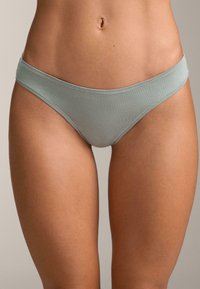 Spodnji del bikinija iz svetlo zelene tkanine z mehko teksturo, nizkim pasom in tanko elastično pasnico. Šivi so vidni ob robovih.