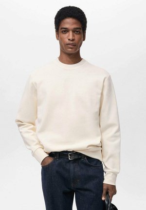 Helles beige Baumwoll-Sweatshirt mit Rundhalsausschnitt, langen Ärmeln und lockerer Passform, kombiniert mit dunkelblauen Jeans.