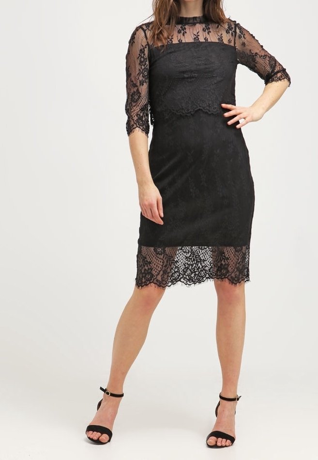 Femme portant une robe noire en dentelle longueur genou avec des manches trois-quarts et des détails en dentelle transparente, associée à des chaussures noires à bride cheville.