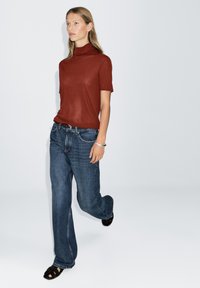Rostrot kurzärmeliges Rollkragenoberteil kombiniert mit entspannt sitzender blauer Jeans. Zusätzliche Details sind ein schwarzer Gürtel und schwarze Sandalen mit Riemchen.