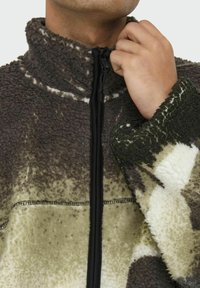 Fleece jacket in brown and green with a high collar and zip closure. Features textured pattern and contrasting black zipper.