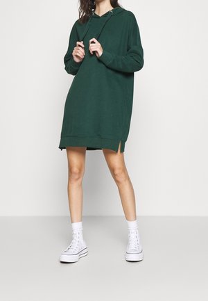 Kvinde iført en mørkegrøn oversized hættetrøjekjole med snore, hvide høje sneakers og hvide sokker mod en ensartet lys baggrund.