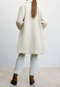 Persona con cabello largo, vistiendo un abrigo texturizado color crema, pantalones claros y botas de tobillo marrones, de pie sobre un suelo liso contra una pared sencilla.