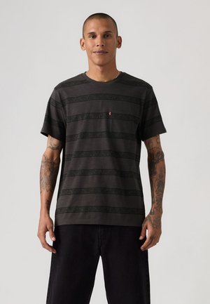 Mann med tatoveringer iført en svart stripete kortermet t-skjorte og svarte bukser, stående mot en enkel lys bakgrunn.