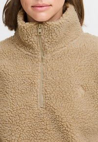 Pullover en polaire beige avec col haut et zip à moitié, doté d'une surface douce et texturée et d'un branding minimaliste sur la poitrine.