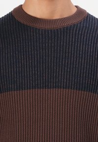 Lähedane vaade ribakotiga kudumisele, mille ülasääre on tume sinine ja alumine osa ning kaelus pruun, kands inimese seljas.