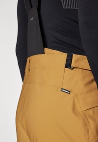 Gule snøscooterbukser med sorte seler, teksturert stoff, sidelommer og logodetalj. Midjebåndet har en glat overflate.