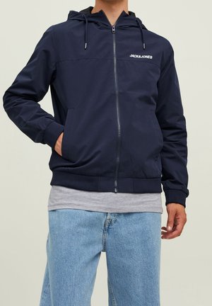 Veste zippée bleu marine avec capuche, poignes élastiques et ourlet inférieur ; dotée d'un logo sur la poitrine et de poches latérales ; tissu léger avec une texture lisse.