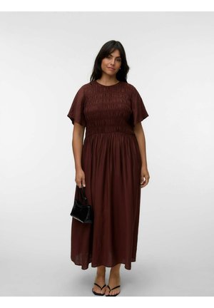 Kvinde med mørkt hår iført en brun smocket midikjole, sorte sandaler og holder en lille sort håndtaske mod en ensfarvet baggrund.