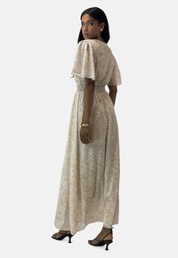 Robe maxi en tissu beige clair avec un motif floral blanc, manches courtes bouffantes, taille smockée et fentes sur les côtés pour faciliter les mouvements.