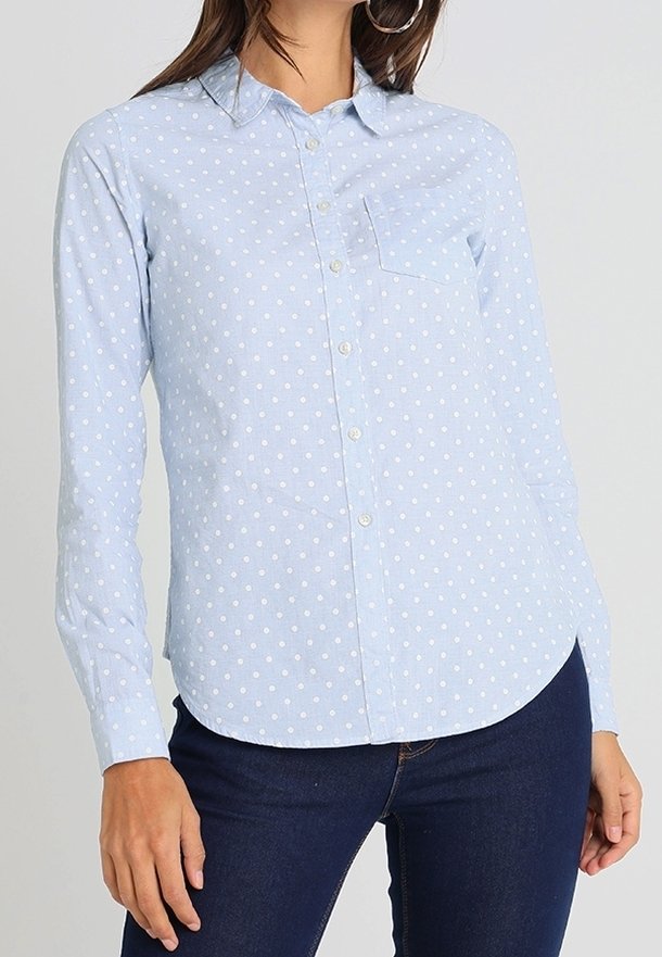 Femme portant une chemise bleu clair à manches longues avec des pois blancs, associée à un jean bleu foncé.