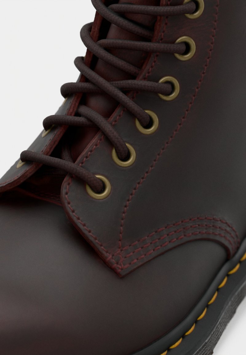 Botte en cuir bordeaux avec des lacets marron foncé, des œillets dorés, des coutures visibles, et une semelle jaune contrastante. Texture lisse, hauteur mi-mollet.