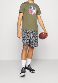 Olivově zelené tričko s logem NFL, šedé kraťasy s abstraktním černým vzorem a černé sportovní boty. Drží hnědý fotbal.