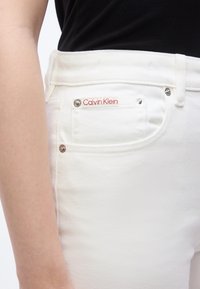 Κοντινό πλάνο στη μέση ενός ατόμου που φοράει λευκά τζιν Calvin Klein και μαύρο τοπ, δείχνοντας την μπροστινή τσέπη με την ετικέτα της μάρκας και τις καρφίτσες.