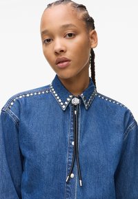 Denim shirt with a buttoned collar, silver stud accents along shoulder seams, and a black cord with silver tips at the neck.