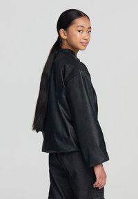 Giacca di pelle nera con vestibilità rilassata, morbido al tatto e maniche larghe, abbinata a pantaloni grigio scuro. Il modello ha i capelli lunghi e lisci.