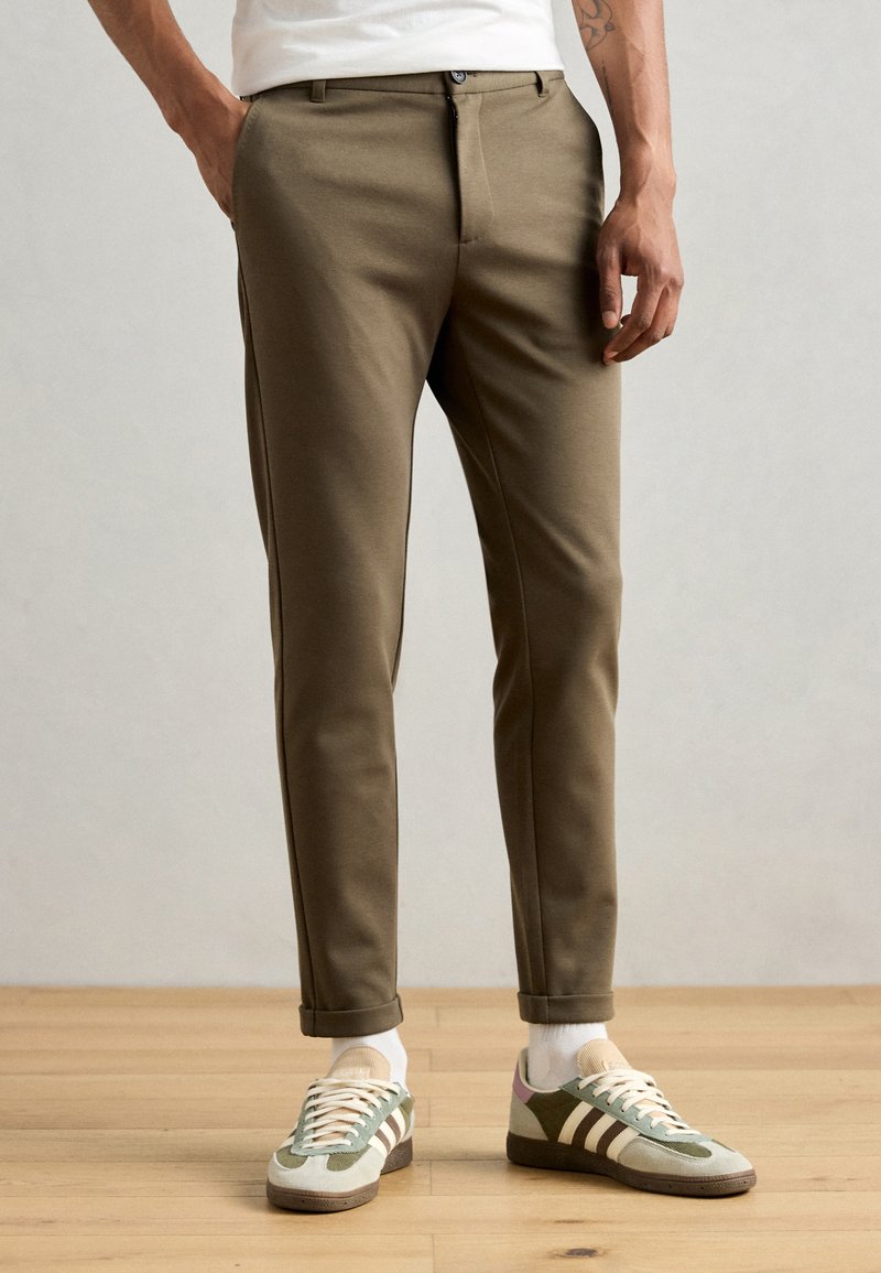 Pantalones khaki ajustados con un corte slim, textura suave y bolsillos laterales. Combinados con zapatillas deportivas verdes y beiges y calcetines blancos hasta el tobillo.