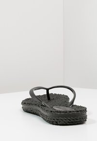 Sandal jari hitam dengan tekstur anyaman, sol datar, dan tali berbentuk V. Solnya memiliki desain anyaman di sepanjang tepi.