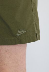 Zelené Nike šortky na noze, s vyšitým logem Nike poblíž lemu, viditelným šitím a detaily bočního švu.