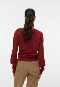 Rode gebreide trui met een V-patroon, ballonmouwen en een aansluitende tailleband, gecombineerd met tan kleurige corduroy broek, gezien van achteren.
