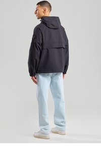 Black hooded jacket with elastic cuffs, loose fit, and a back vent. Worn with light blue jeans and white sneakers.
