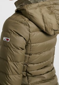 Chaqueta de plumas verde oliva con capucha, caracterizada por paneles horizontales cosidos y un pequeño parche con logotipo en la parte superior del brazo. Textura suave y brillante.