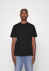 T-shirt in cotone nero con maniche corte e collo rotondo; presenta un piccolo logo sul lato sinistro del petto; abbinato a jeans blu.