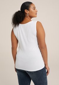 Top blanco sin mangas con cuello redondo y textura suave. Con un dobladillo trasero ligeramente más largo. Combinado con jeans azul oscuro.