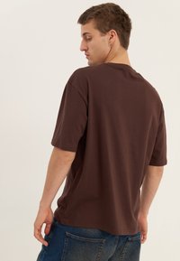 Ung mann med kort brunt hår iført en løs, mørk brun T-skjorte og blå jeans, som ser over skulderen mot en enkel bakgrunn.