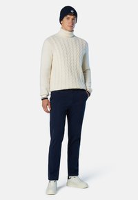 Cremfarbener Zopfmuster-Pullover mit hohem Kragen, kombiniert mit navyfarbenen Chinos und weißen Turnschuhen. Das Modell trägt außerdem eine navyfarbene Mütze mit Logo-Details.