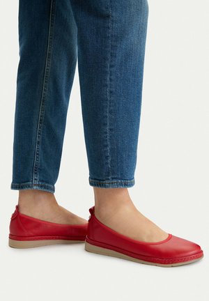 Füße, die rote Leder-Slipper tragen, kombiniert mit hochgekrempelten blauen Jeans, vor weißem Hintergrund.