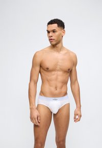 Αθλητικός άνδρας με κοντά σγουρά μαλλιά που φοράει λευκά εσώρουχα Calvin Klein, στέκεται και κοιτάζει προς το πλάι μπροστά σε απλό ανοιχτό φόντο.