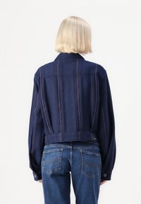 Σκούρο μπλε κοντό σακάκι με κατακόρυφες ρίγες, κουμπωμένα μανίκια και γιακά. Φοριέται πάνω από μπλε τζιν παντελόνι; υφασμάτινη επιφάνεια με λεπτομέρειες ραφής.