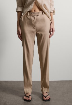 Frau trägt beige hoch taillierte, gerade geschnittene Hosen und schwarze Sandalen mit quadratischer Spitze, steht vor einem einfarbigen hellen Wandhintergrund.