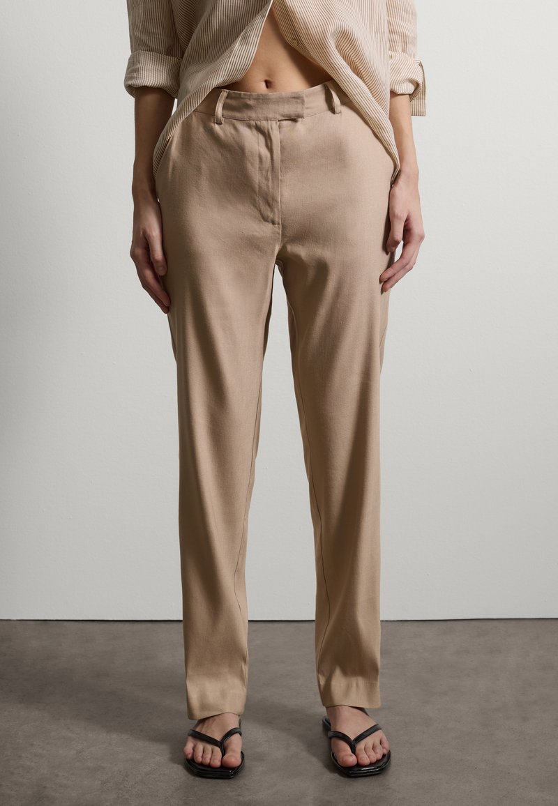 Frau trägt beige hoch taillierte, gerade geschnittene Hosen und schwarze Sandalen mit quadratischer Spitze, steht vor einem einfarbigen hellen Wandhintergrund.