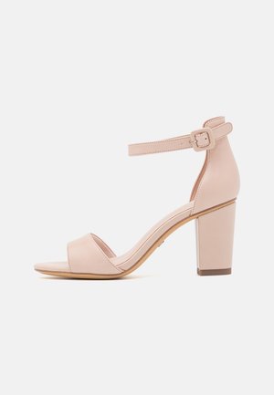 Beige sandal med åben tå, ankelrem med spænde, blokhæl og glat finish på en hvid baggrund.