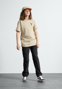 Beige cotton T-shirt with logo, black pants, and black and white sneakers. The cap is multicolored with a flat brim.