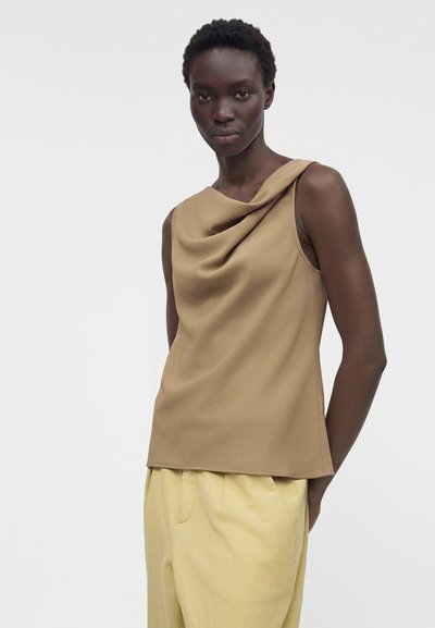 Γυναίκα με κοντά σγουρά μαλλιά που φοράει ένα αμάνικο μπεζ τοπ με πλισέ και ανοιχτόκκινο κίτρινο παντελόνι, στέκεται μπροστά σε ένα απλό γκρι φόντο.