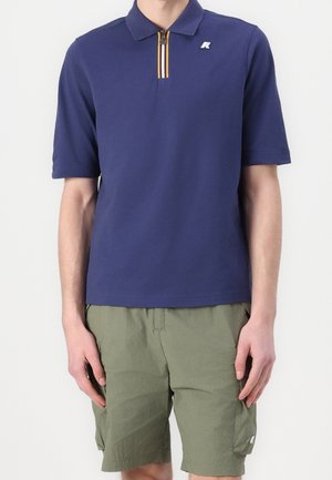 Homme portant un polo bleu marine à manches courtes avec une fermeture éclair rayée et un short cargo vert olive sur fond blanc.