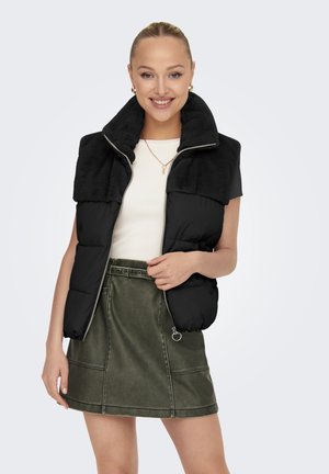 Gilet nero imbottito con collo in pelliccia sintetica, zip argentata e tasche laterali, abbinato a una gonna verde in ecopelle e una maglietta bianca.