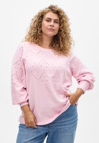 Lichtroze trui met een diamant-lace patroon, ruime mouwen en geribbelde manchetten. Gepaard met losse spijkerbroeken.