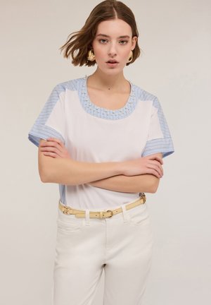 Witte t-shirt met lichtblauwe gestreepte mouwen, versierd met een bewerkte halslijn. Gecombineerd met lichtgekleurde broek en een beige riem.